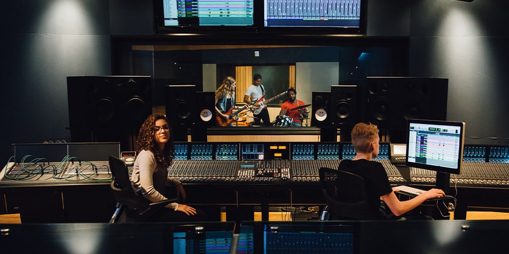 Woman and man sitting behind console in studio with a band behind glass.