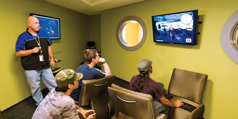 Three men in a yellow room watching one man play video game on tv screen.