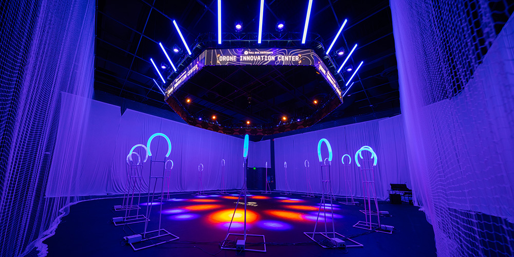 Full Sail’s Drone Innovation Center lit in blue and purple, with safety nets and glowing racing gates arranged inside a large indoor flight zone.