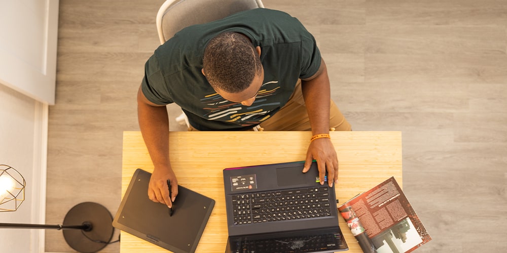 A photograph taken above of a man sitting at a desk with a laptop and drawing on a Wacom tablet.