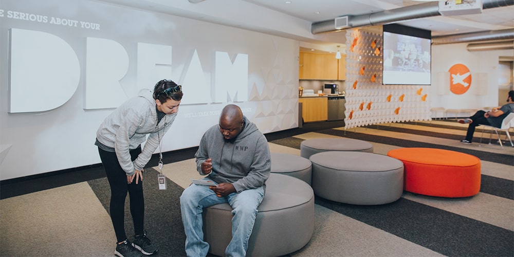 A female employee leaning over while talking to a male who is sitting on an ottoman in a room with light and dark grey striped flooring and the word DREAM projected on the wall.