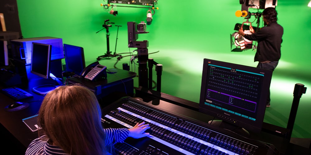 Woman behind console looking at man adjusting a light on a green screen.