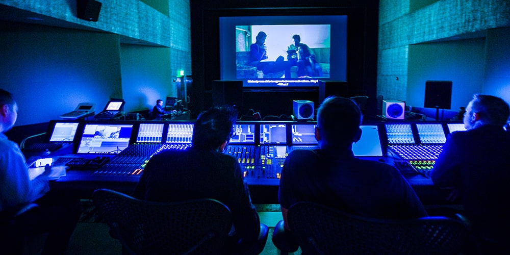 Four men behind console in dubbing stage watching film on big screen.